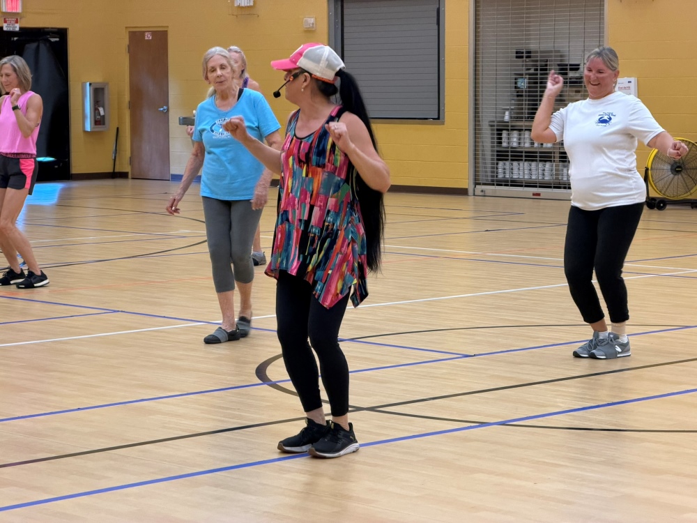 A group of women spread out and dancing along with a woman in the foreground, who has a hat and a microphone